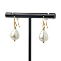 The Pearl Earrings in Gold feature elegant teardrop-shaped white pearls on gold hooks, displayed on a black stand against a white background for a timeless appearance.