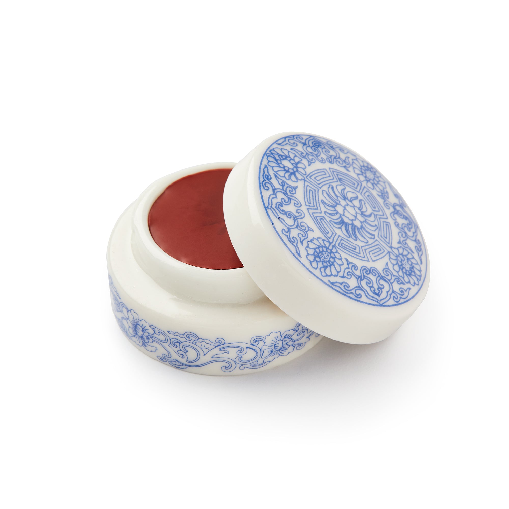 The Crème Rouge comes in a round white porcelain pot with intricate blue designs. Its partially open lid reveals the cream rouge inside, and features a detailed circular pattern.