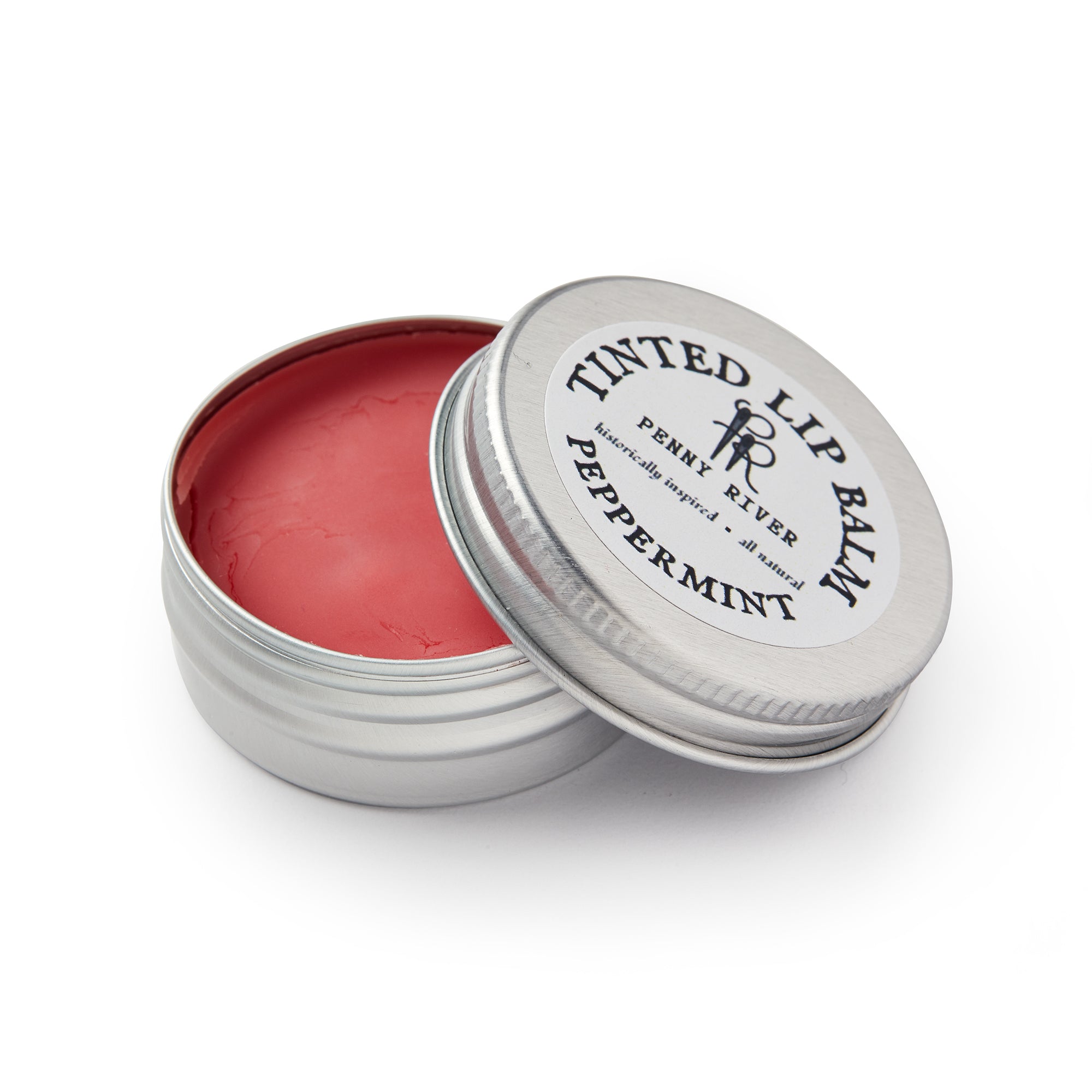 A round, silver tin of moisturizing Lip Balm in peppermint, its lid partly open to reveal smooth pink balm. The lid reads “Tinted Lip Balm, Penny River, Peppermint.”.