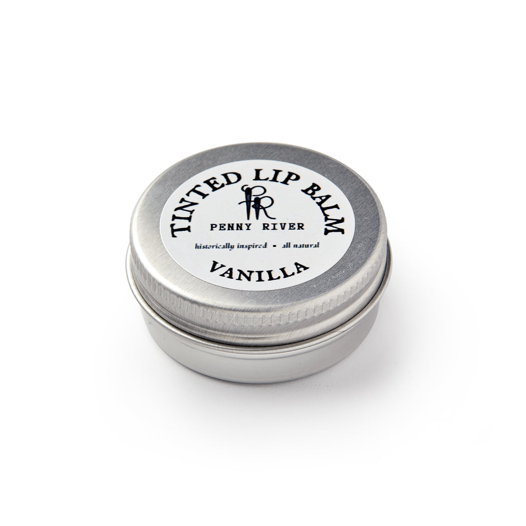 A small, round metal tin labeled Lip Balm Vanilla by Penny River sits closed on a white background. The lid features the text "historically inspired • all natural." This is a moisturizing tinted lip balm.