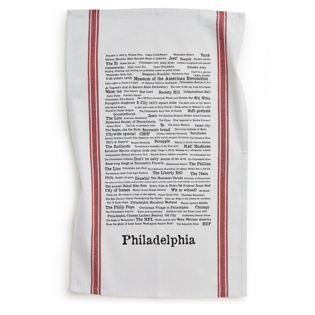 The Philadelphia Tea Towel is white with red stripes on the sides, handprinted with black text listing famous local trivia, foods, and cultural references above “Philadelphia.”.