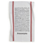 The Philadelphia Tea Towel is white with red stripes on the sides, handprinted with black text listing famous local trivia, foods, and cultural references above “Philadelphia.”.