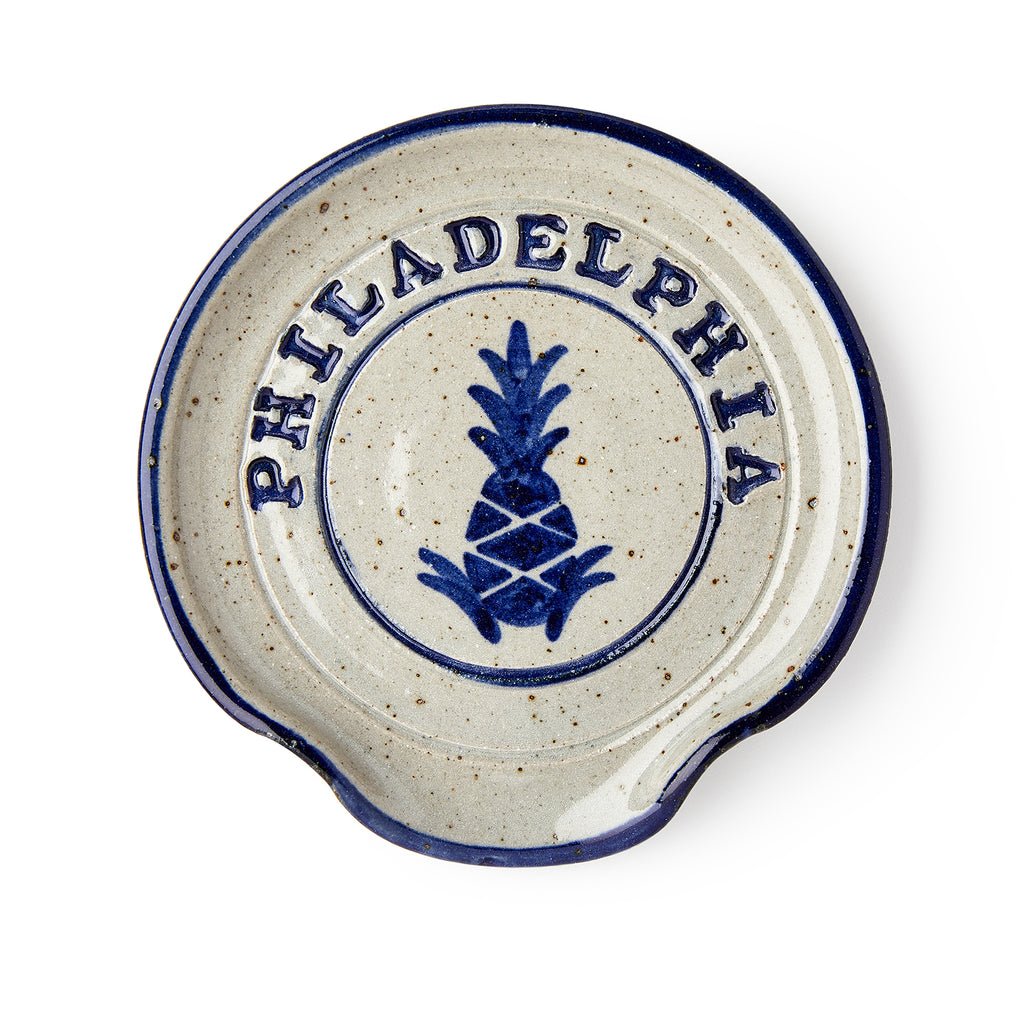 The Philadelphia Stoneware Spoon Rest is a round dish with a blue rim, a blue pineapple motif, and "PHILADELPHIA" in capitals above it. It features a small curved notch on one side for easy spoon resting.
