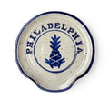 The Philadelphia Stoneware Spoon Rest is a round dish with a blue rim, a blue pineapple motif, and "PHILADELPHIA" in capitals above it. It features a small curved notch on one side for easy spoon resting.