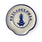 The Philadelphia Stoneware Spoon Rest is a round dish with a blue rim, a blue pineapple motif, and "PHILADELPHIA" in capitals above it. It features a small curved notch on one side for easy spoon resting.