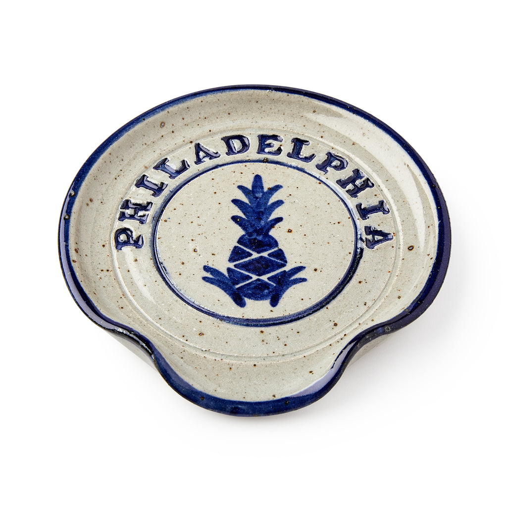 The Philadelphia Stoneware Spoon Rest features a round design with a blue rim, a central blue pineapple motif, bold "PHILADELPHIA" lettering on top, and a side notch for easy utensil placement.