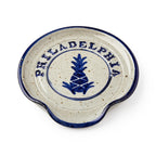 The Philadelphia Stoneware Spoon Rest features a round design with a blue rim, a central blue pineapple motif, bold "PHILADELPHIA" lettering on top, and a side notch for easy utensil placement.