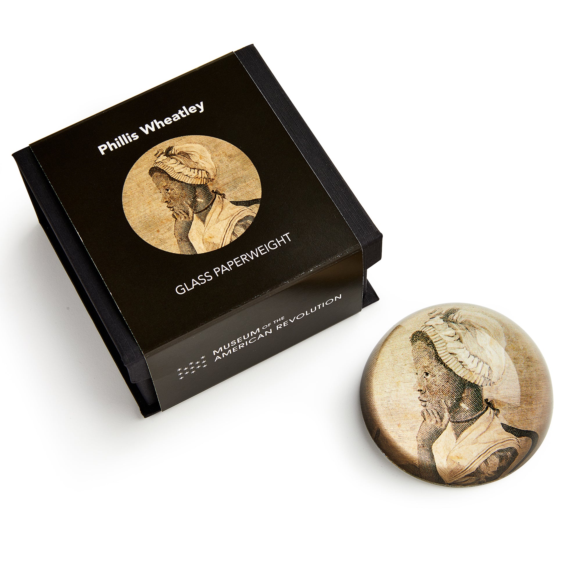 A round glass paperweight featuring an illustration of African American poet Phillis Wheatley, shown beside its black box labeled "Phillis Wheatley Glass Paperweight" from the Museum of the American Revolution.
