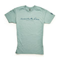 The Remember the Ladies T-Shirt is light green with short sleeves, featuring “Remember the ladies – Abigail Adams” in navy cursive on the chest and a small logo on the left sleeve. Shirt shown laid flat on a white background.