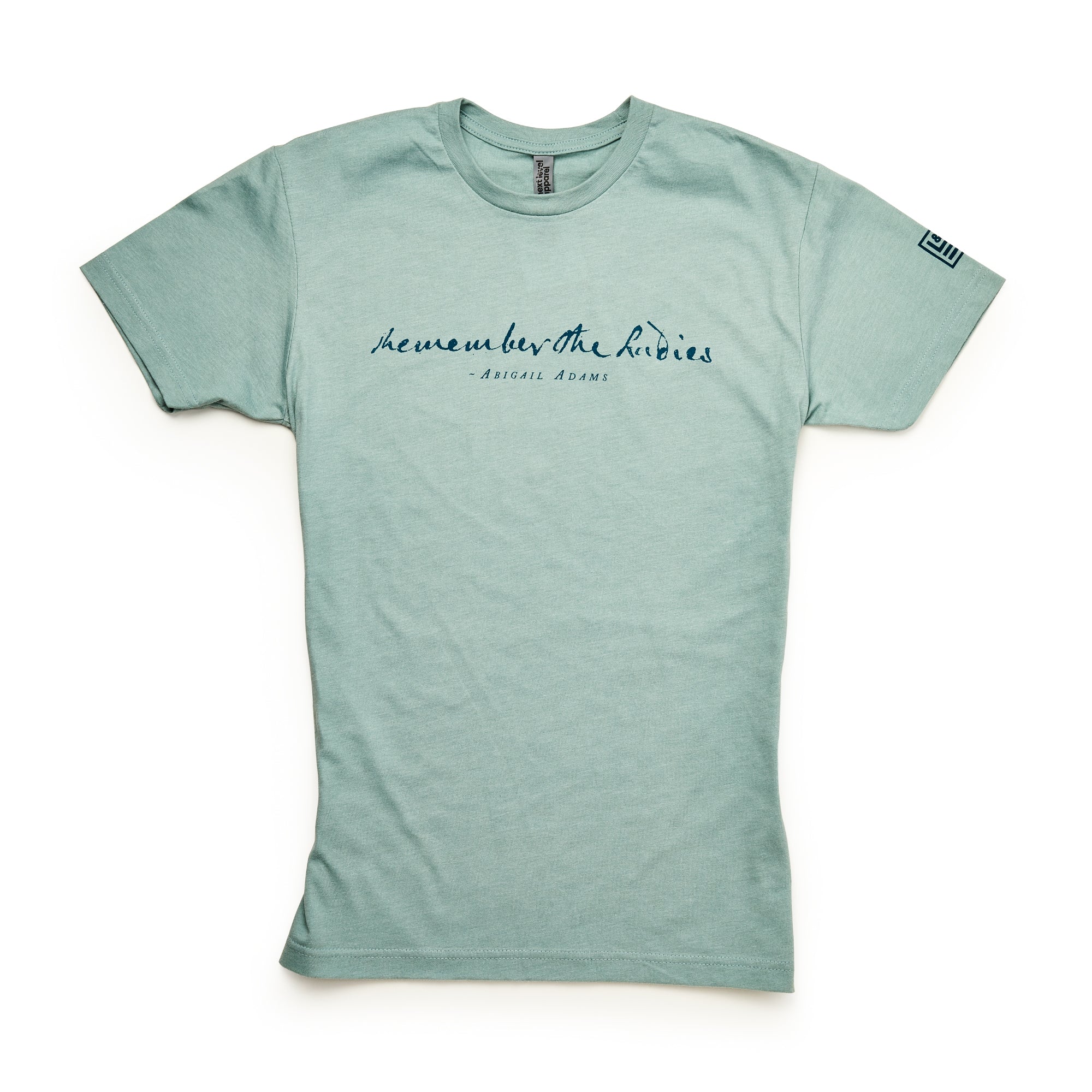 The Remember the Ladies T-Shirt is light green with short sleeves, featuring “Remember the ladies – Abigail Adams” in navy cursive on the chest and a small logo on the left sleeve. Shirt shown laid flat on a white background.