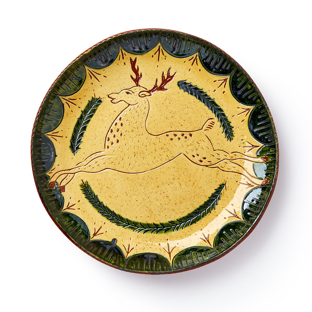 The Deer Plate is a handcrafted ceramic piece with a yellow background, leaping deer with red antlers and brown spots, green leaf border accents, and two curved branches beneath for classic folk pottery charm.
