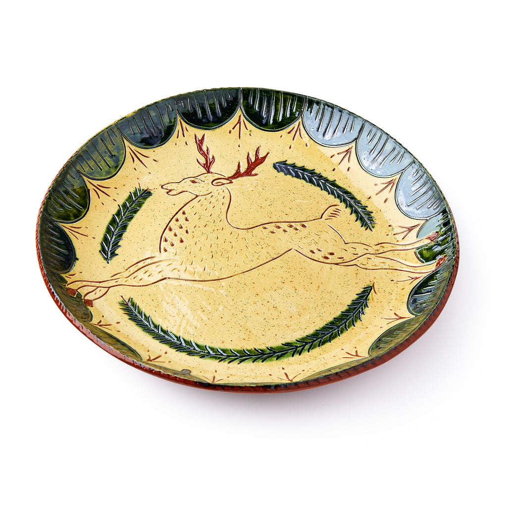 The Deer Plate is a handcrafted redware sgraffito piece featuring a hand-painted leaping deer at the center, green leaf motifs, and a scalloped green and yellow border, inspired by traditional folk pottery.