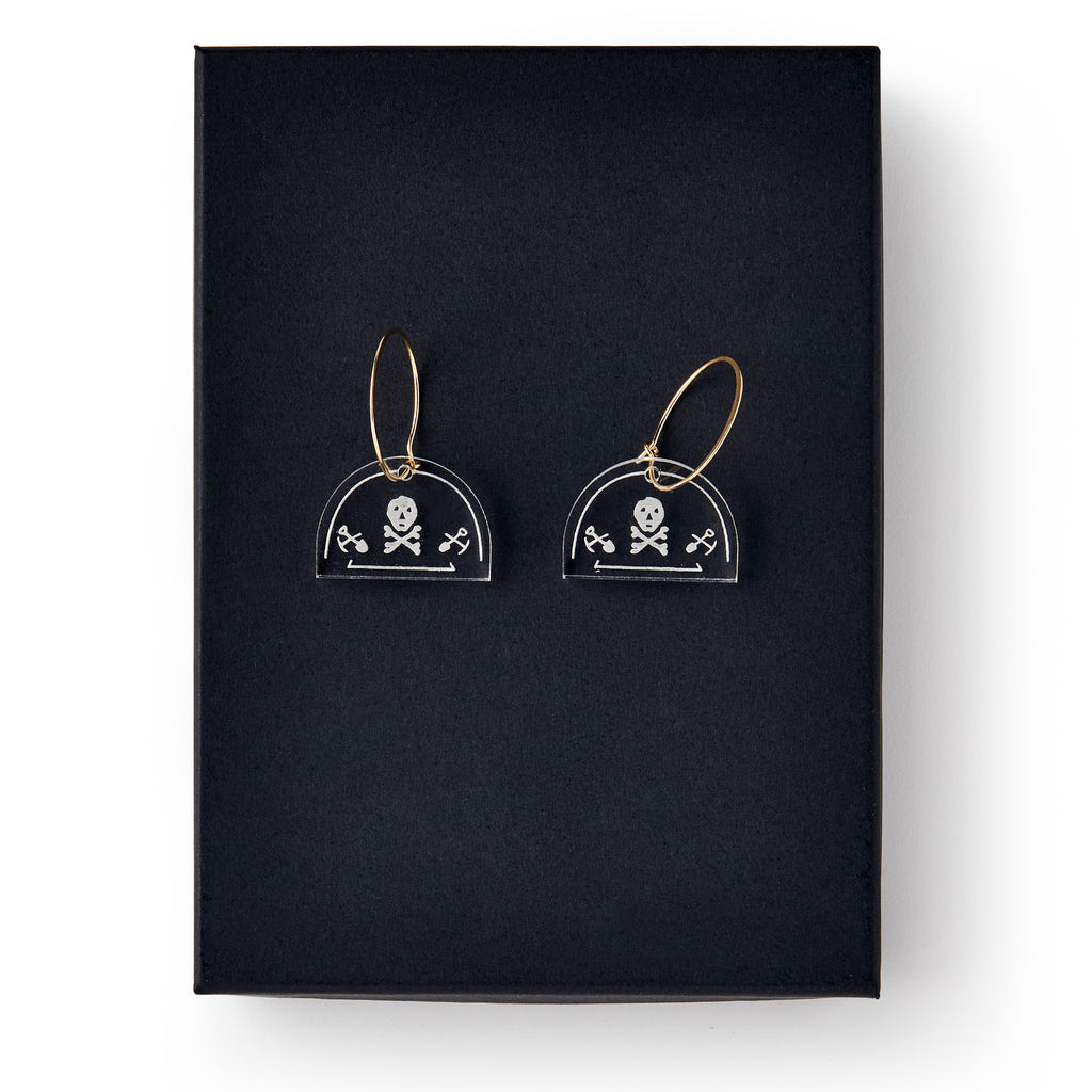 Skull and Crossbone Earrings: Clear acrylic earrings with gold hoops, each featuring a white skull-and-crossbones design, displayed on black—ideal for fans of historical or Stamp Act-inspired jewelry.