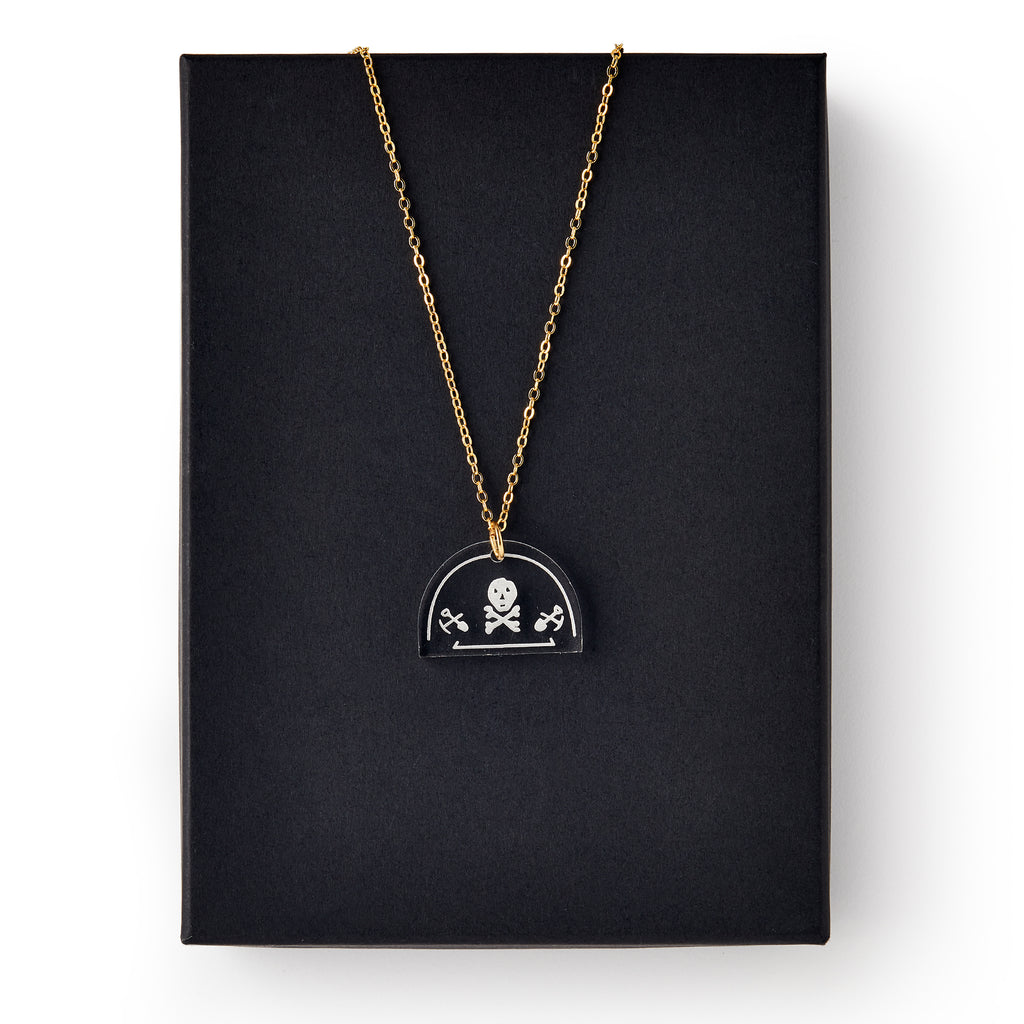 The Skull and Crossbones Necklace features a gold chain and black pendant, inspired by Stamp Act jewelry and colonial resistance symbols, displayed on a plain black rectangular box.