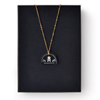 The Skull and Crossbones Necklace features a gold chain and black pendant, inspired by Stamp Act jewelry and colonial resistance symbols, displayed on a plain black rectangular box.