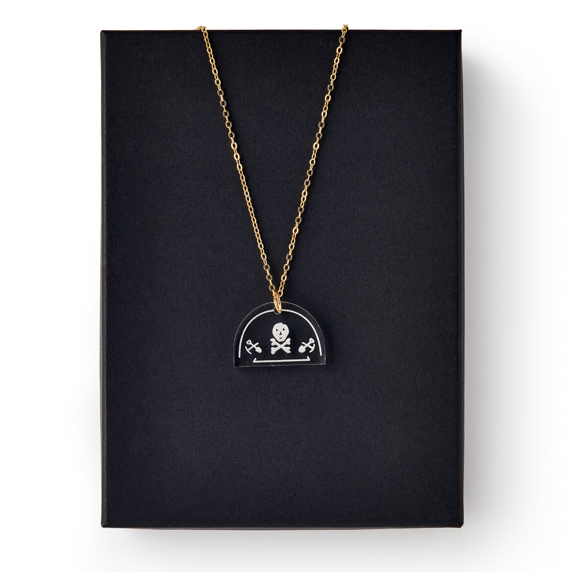 The Skull and Crossbones Necklace features a gold chain and black pendant, inspired by Stamp Act jewelry and colonial resistance symbols, displayed on a plain black rectangular box.