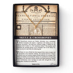 The Skull and Crossbones Necklace comes in a display box with a replica Pennsylvania Journal featuring the skull and crossbones image, colonial resistance details, and a gold necklace pendant shaped like a skull and crossbones.