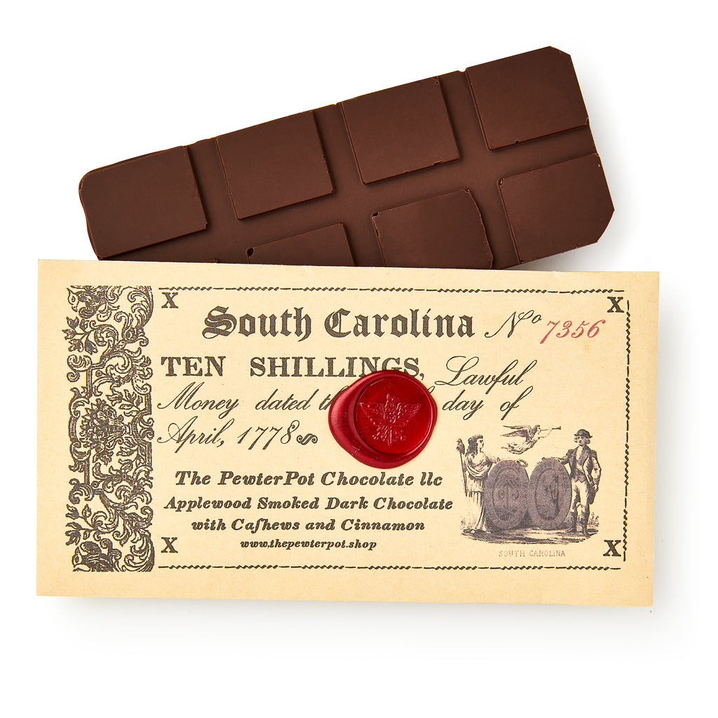 The South Carolina Chocolate Bar, made with smoked chocolate, rests on a vintage-inspired ten shilling note adorned with a red wax seal, old-style text, and illustrations of barrels and two men.