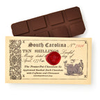 The South Carolina Chocolate Bar, made with smoked chocolate, rests on a vintage-inspired ten shilling note adorned with a red wax seal, old-style text, and illustrations of barrels and two men.