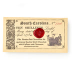 A vintage-style South Carolina Ten Shillings note with ornate borders, a red wax seal, and illustrations sits above text promoting the South Carolina Chocolate Bar—applewood smoked chocolate with cashews and cinnamon.