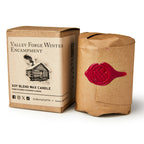 A brown box labeled Valley Forge Candle stands beside a candle wrapped in brown paper, sealed with red wax and a decorative stamp—both evoking a rustic, handcrafted look ideal for adding historical ambiance.