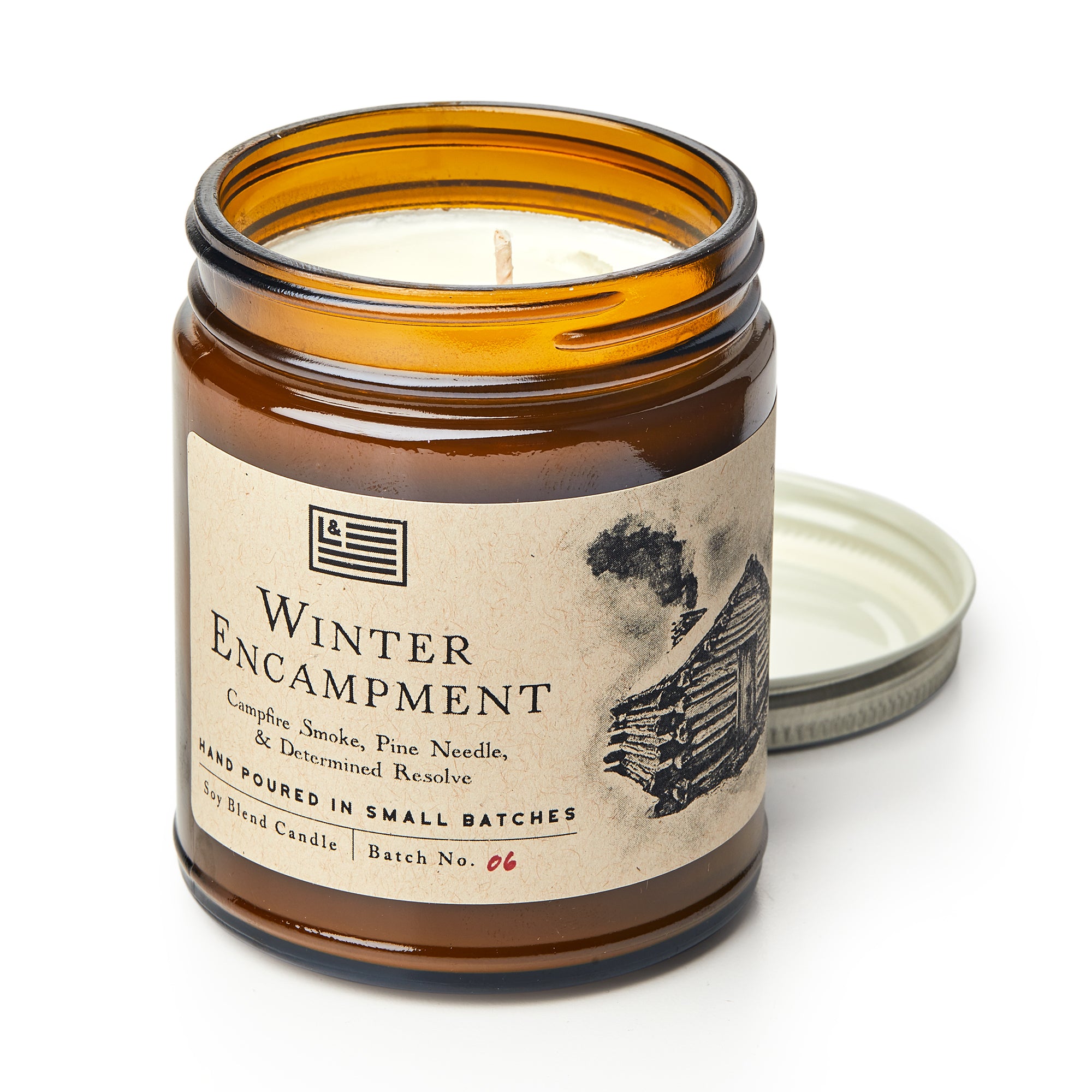 The Valley Forge Candle, labeled "Winter Encampment," comes in an amber glass jar and features a log cabin with smoke. Behind the jar rests the lid. Notes of campfire smoke and pine needle create a warm, historical ambiance.