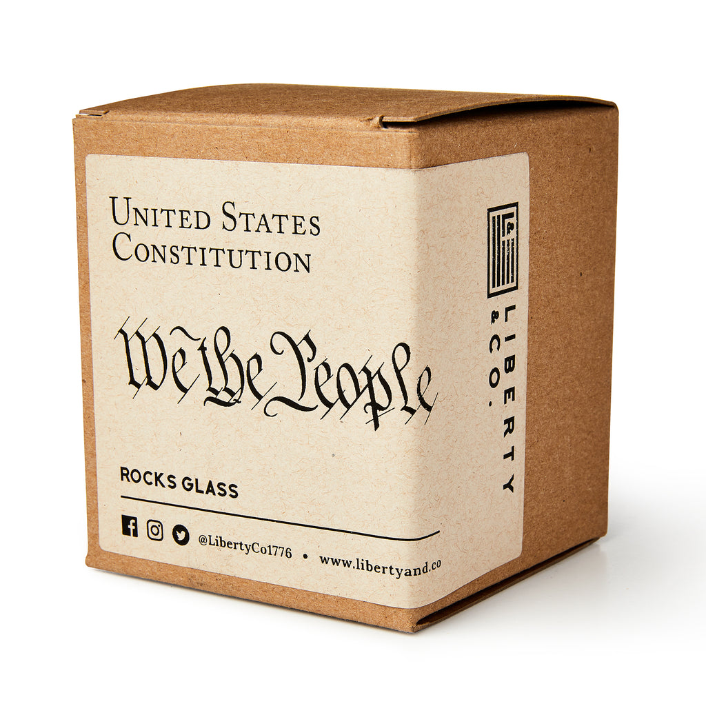 A brown cardboard box labeled "We The People Whiskey Glass" from Liberty & Co., containing the whiskey glass, with social media icons and the company website on the packaging.