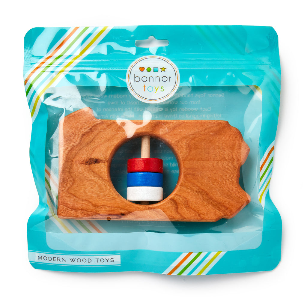 The Pennsylvania State Wooden Rattle is shaped like Pennsylvania, features a spinning red, white, and blue cylinder in the center, and comes in a teal Bannor Toys bag.