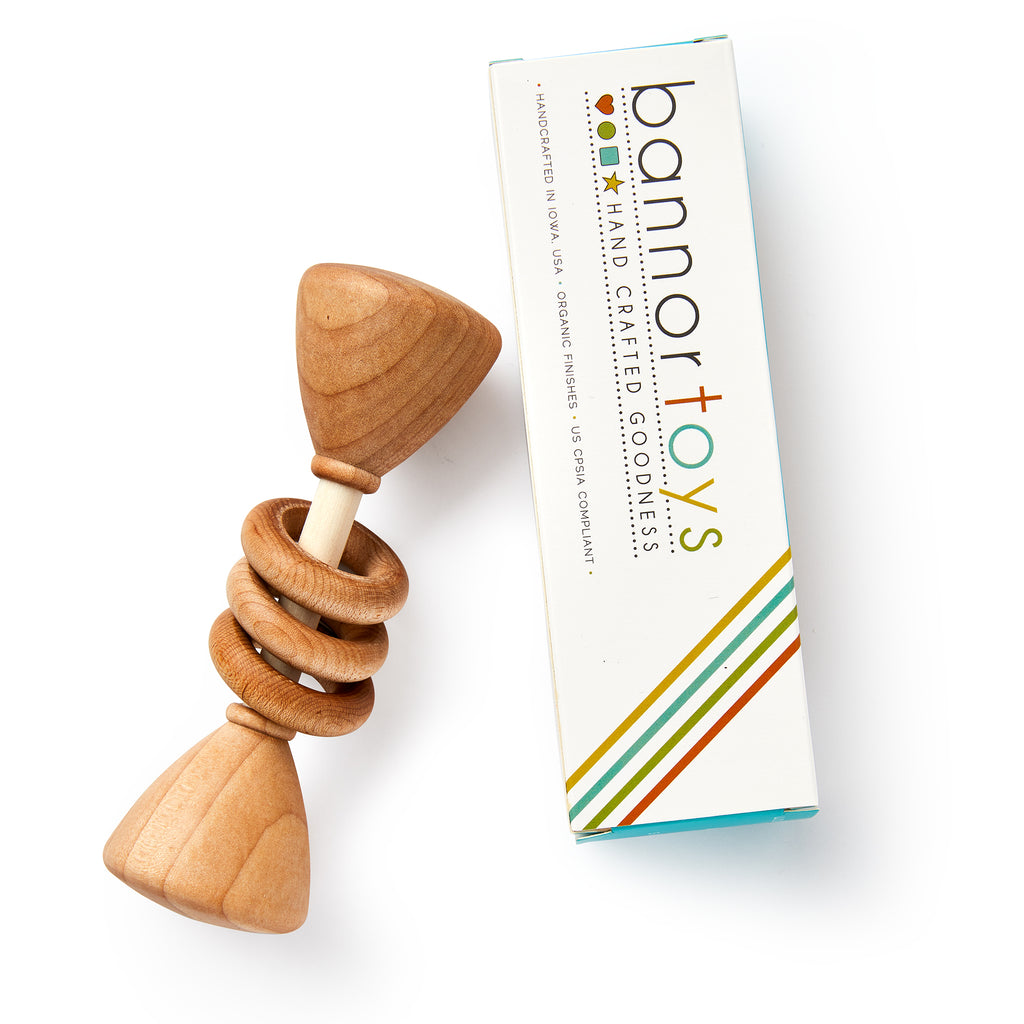 A Wooden Baby Rattle with three rings rests beside a rectangular box labeled Bannor Toys, decorated with colorful stripes and text that highlights it as a handcrafted natural baby toy made with organic finishes.