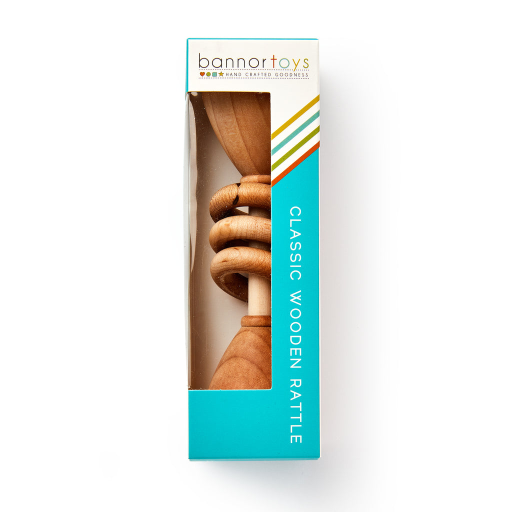 A Wooden Baby Rattle is shown in a turquoise and white box with a window revealing the natural wood toy. The box says "Classic Wooden Rattle" and "Hand Crafted Goodness" — the perfect natural baby toy.