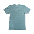 A light blue short-sleeve Remember the Ladies T-Shirt (MoAR) featuring “remember the lilies” and “Jasmin Arora” in delicate cursive across the chest, inspired by iconic women’s rights figures. The commemorative tee is displayed flat on a white background.