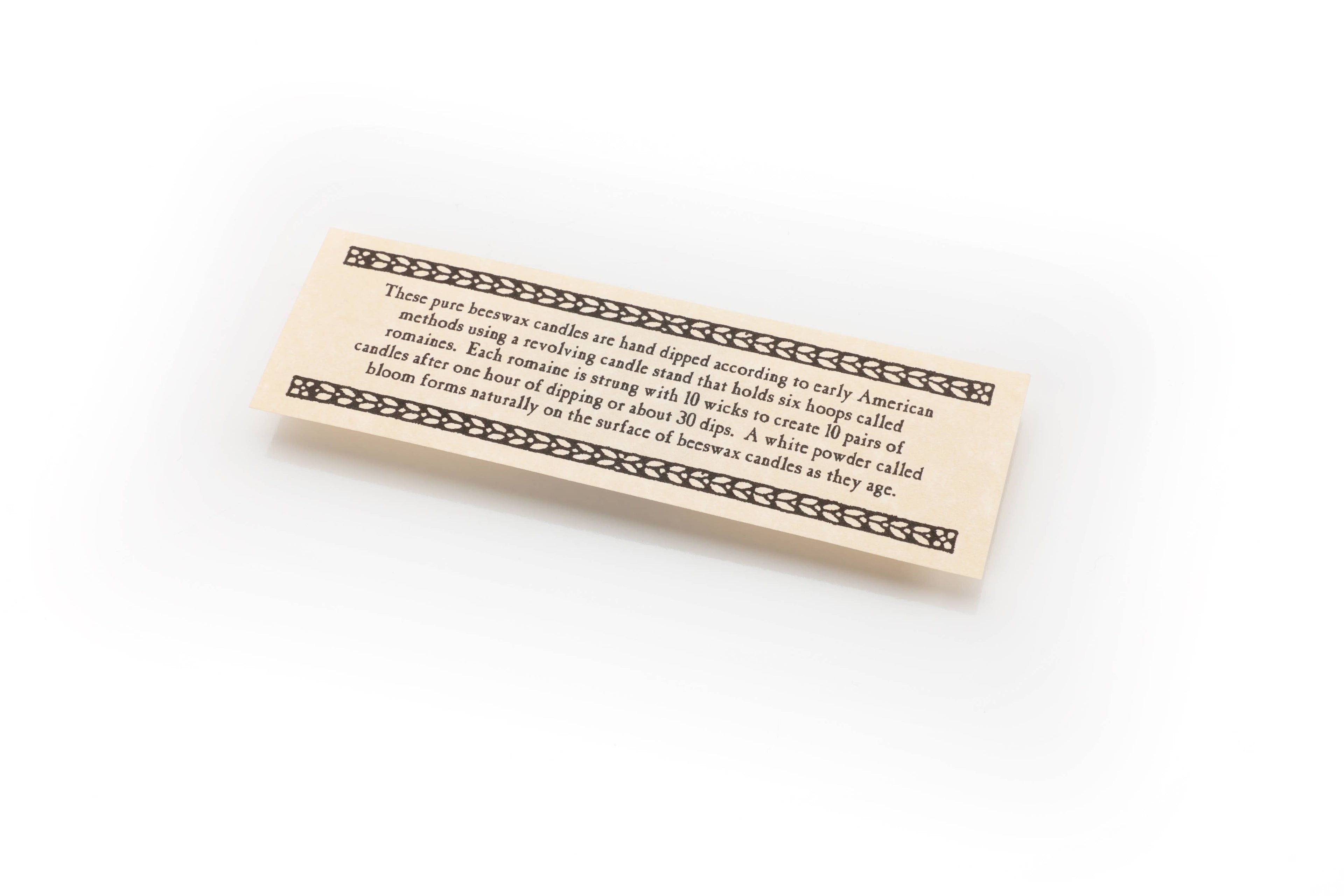 A cream-colored rectangular card with a decorative border displays printed info about Beeswax Taper Candle, hand-dipped using early American methods. The card rests on a white background.