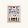 The Colonial Sampler Counted Cross Stitch Kit creates a framed sampler with sheep, a red heart, blue alphabet, two gold stars, “1776” in red, trees, a house, and a red patterned border in traditional American style.