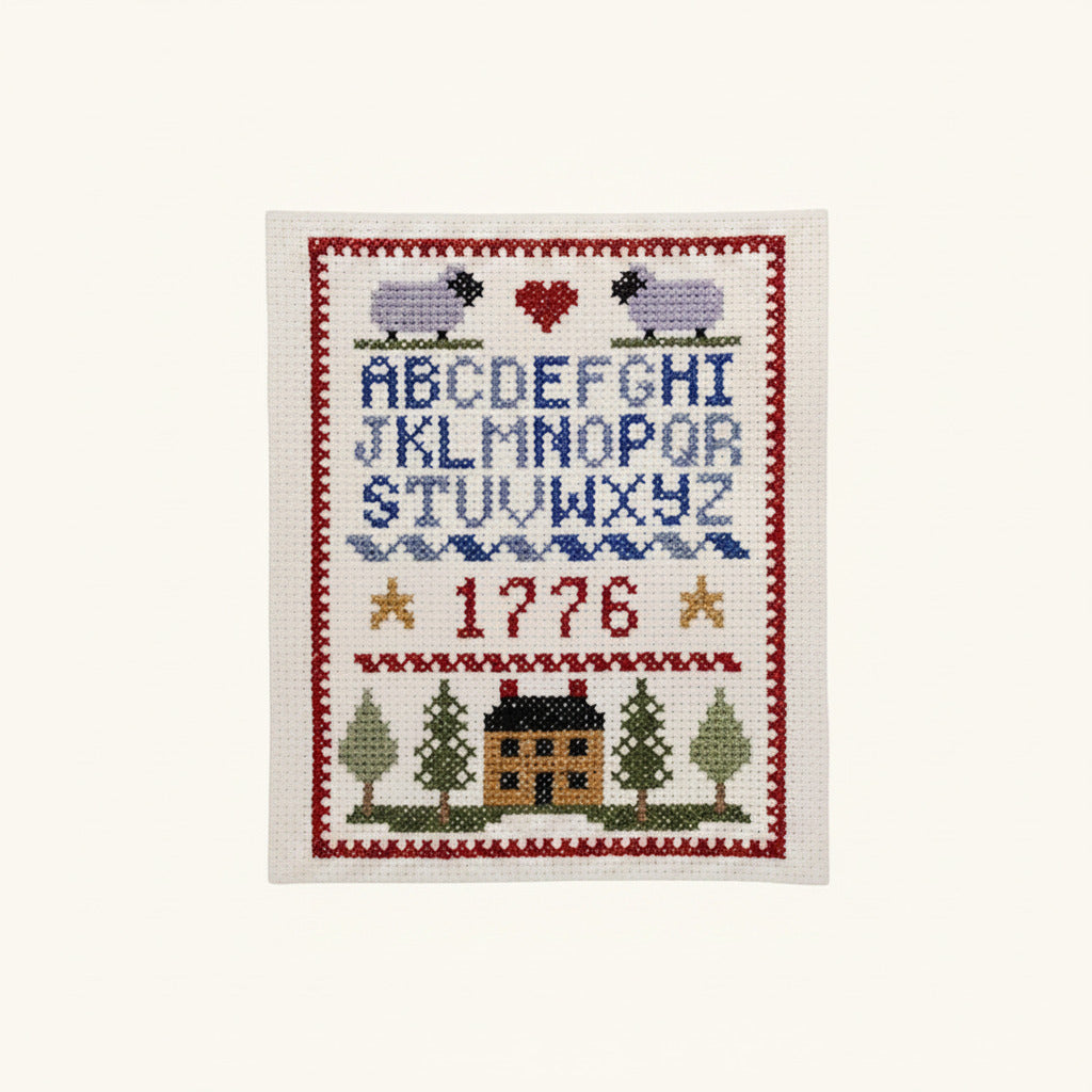 The Colonial Sampler Counted Cross Stitch Kit creates a framed sampler with sheep, a red heart, blue alphabet, two gold stars, “1776” in red, trees, a house, and a red patterned border in traditional American style.