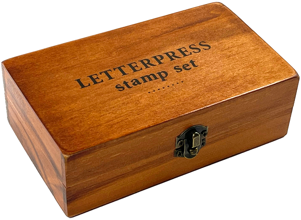 The Letterpress Stamp Set comes in a rectangular wooden box with a black-lettered lid and latch, containing rubber-tipped wooden stamps ideal for creative projects.