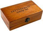 The Letterpress Stamp Set comes in a rectangular wooden box with a black-lettered lid and latch, containing rubber-tipped wooden stamps ideal for creative projects.