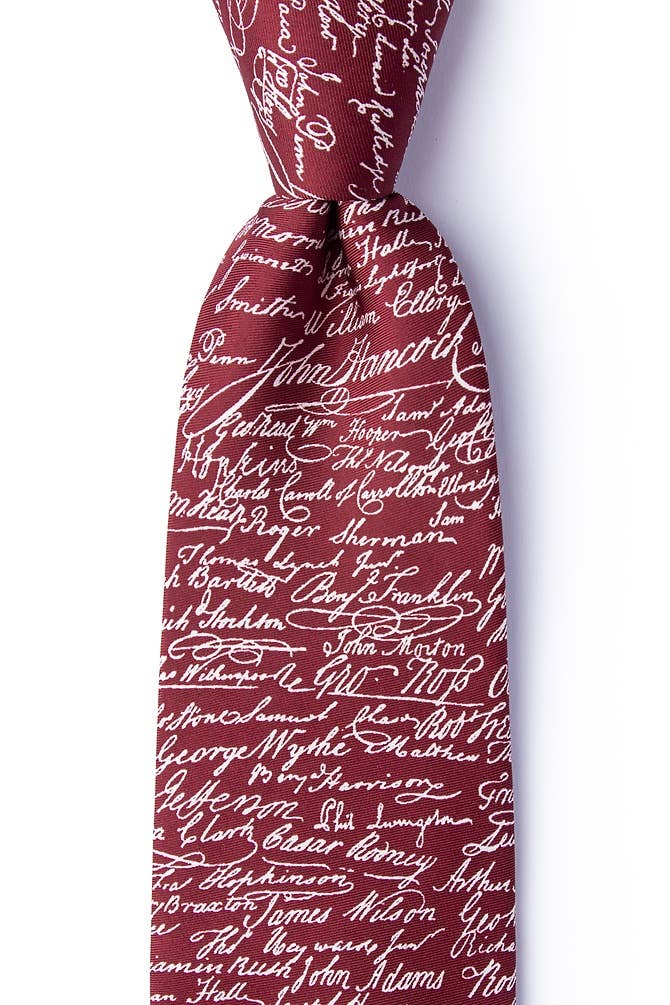 A close-up of the Declaration of Independence Signers Silk Tie showcases its maroon silk fabric adorned with white cursive signatures, forming a distinctive all-over pattern. Knotted and set against a plain white background, this tie makes a perfect gift for history enthusiasts or a stylish accessory for patriotic celebrations.