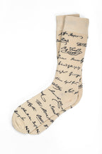 Introducing the Declaration Signers Crew Socks: beige crew socks featuring a repeating pattern of black cursive signatures reminiscent of the historic Declaration of Independence. These stylish socks add a subtle touch of patriotism to your wardrobe, with their signature motif set against a classic beige background. Presented here laid flat on a white backdrop.