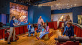 A museum guide addresses visitors on benches within a ship exhibit at the Museum of the American Revolution, surrounded by cannons and historic decor.