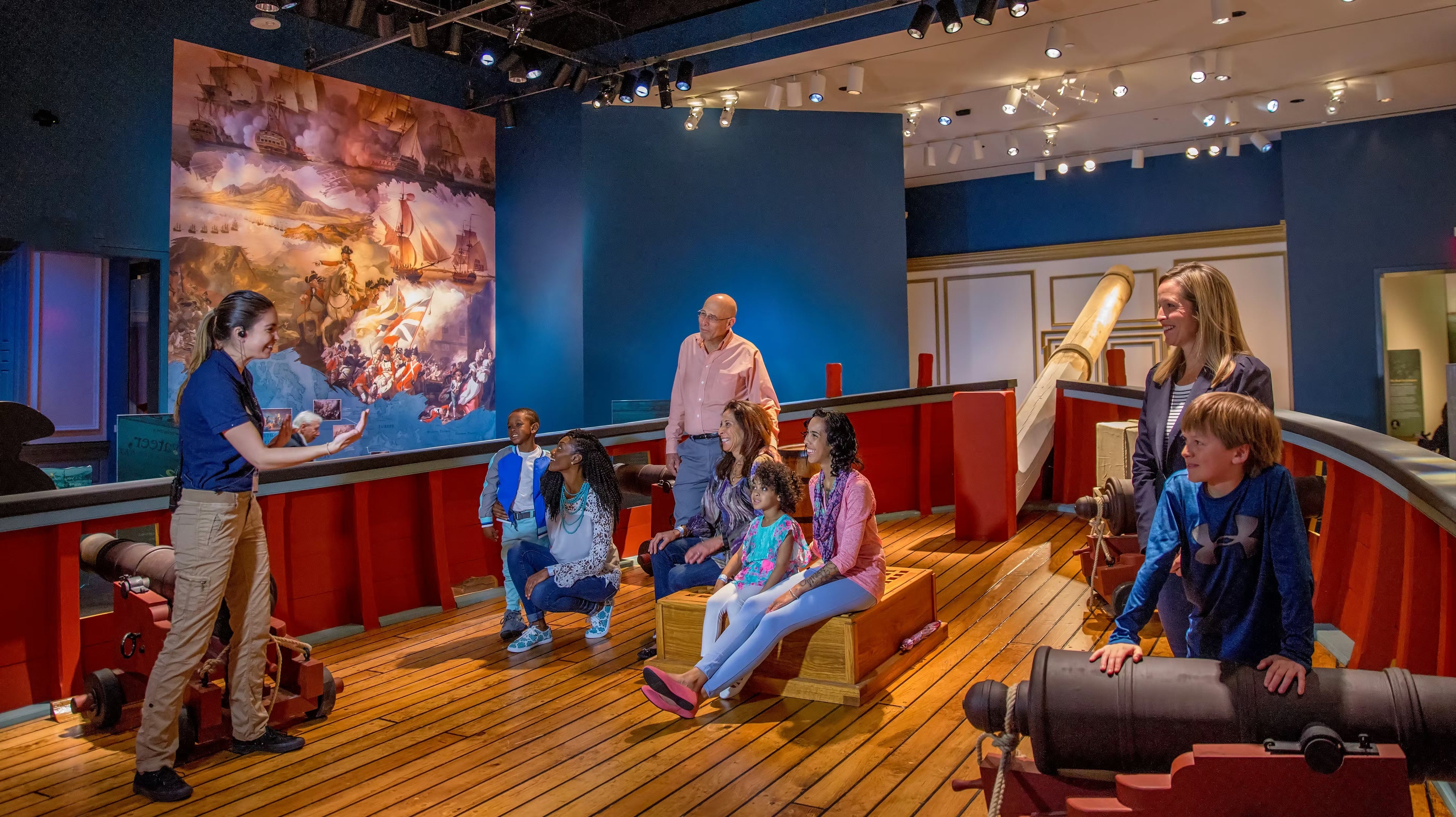 A museum guide addresses visitors on benches within a ship exhibit at the Museum of the American Revolution, surrounded by cannons and historic decor.